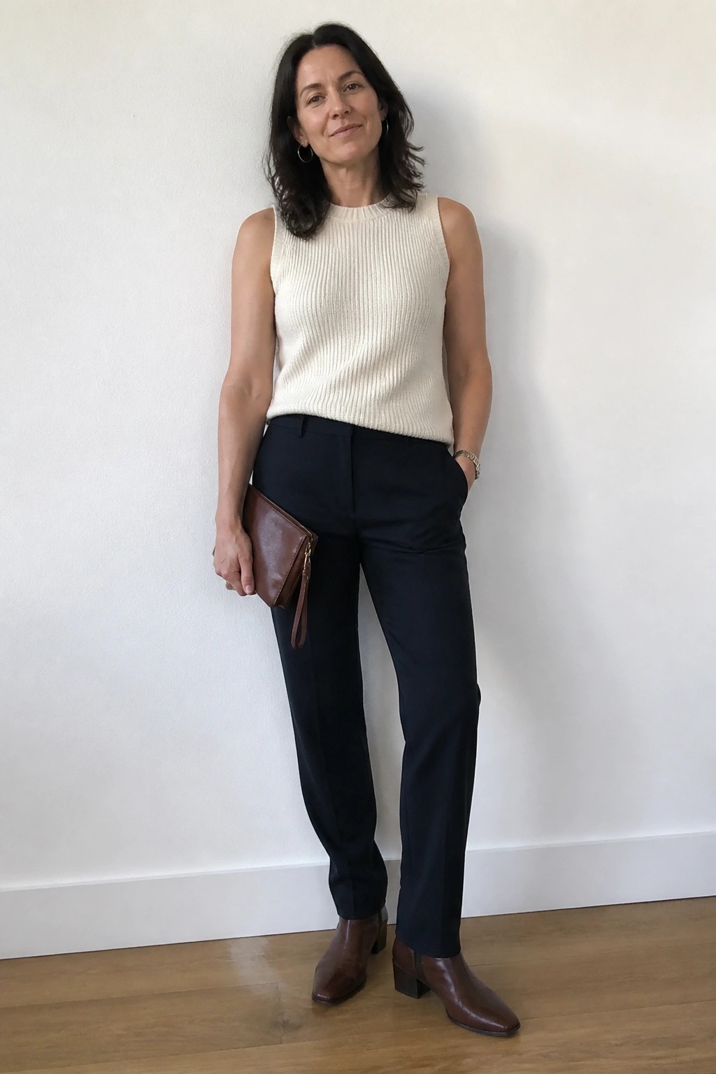 Woman in cream sleeveless knit top, navy pants, brown boots holding clutch