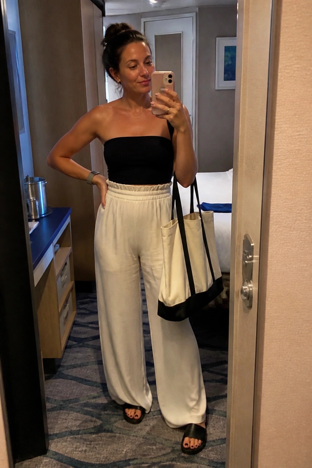 Woman in black strapless bandeau top and white wide leg pants holding a tote bag while taking a mirror selfie in a hotel room.