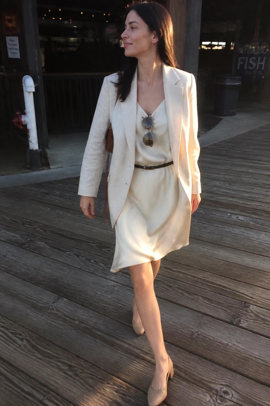 Woman in white blazer over satin dress walking on wooden boardwalk.