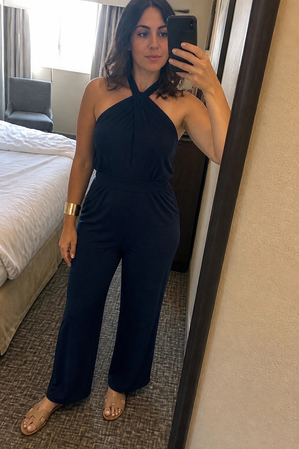 Woman in navy halter jumpsuit takes mirror selfie in hotel room wearing beige sandals