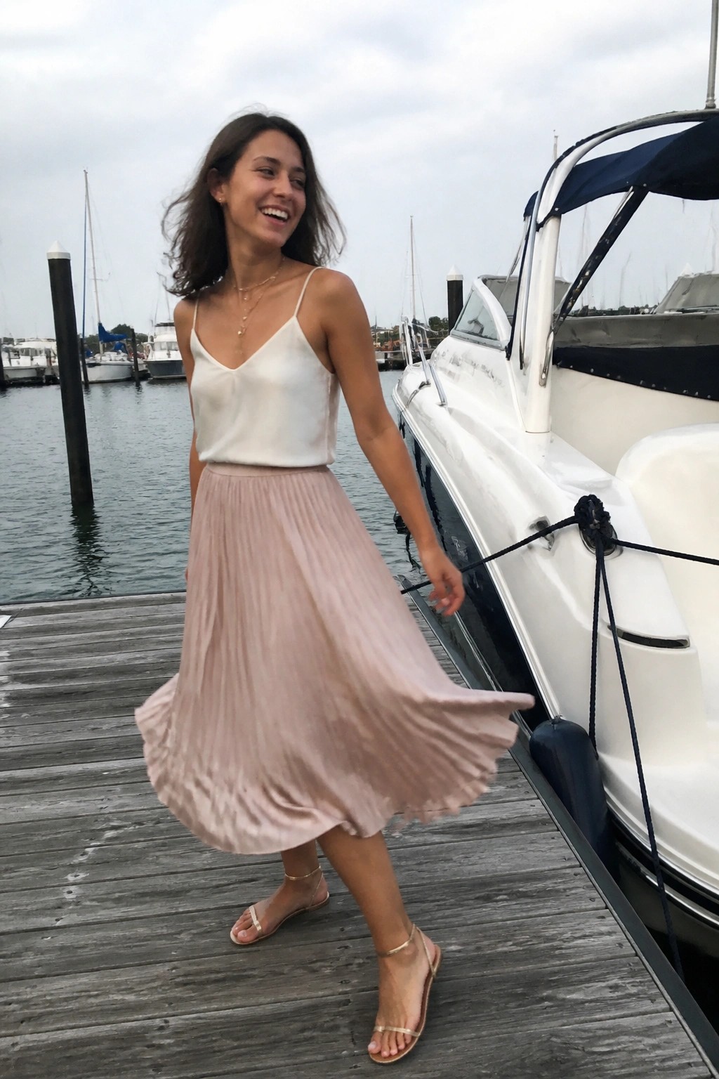 A woman wearing a white camisole top and a light pink pleated midi skirt with gold sandals and jewelry standing on a wooden dock next to a white boat.