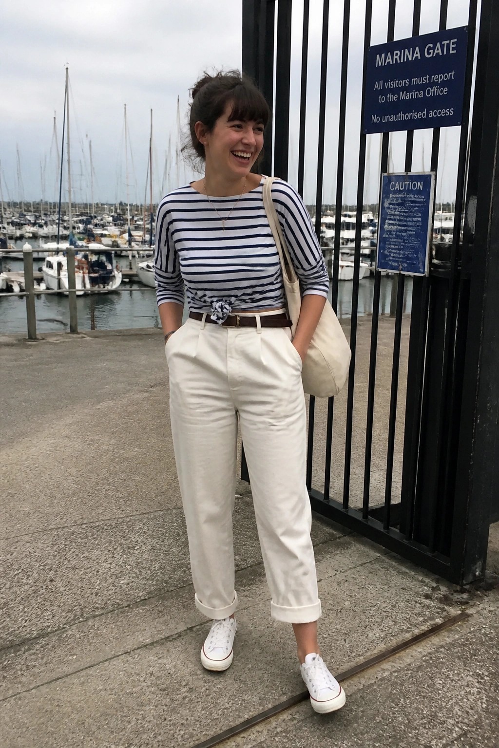 Woman wearing navy and white striped three quarter sleeve top tied at the waist, high waisted white cuffed pants with brown belt, white canvas sneakers, and beige tote bag standing near a marina gate.
