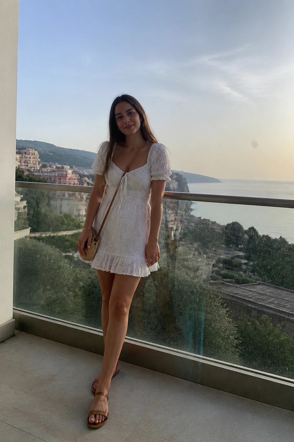 Woman in white eyelet mini dress on balcony overlooking coastal town and sea