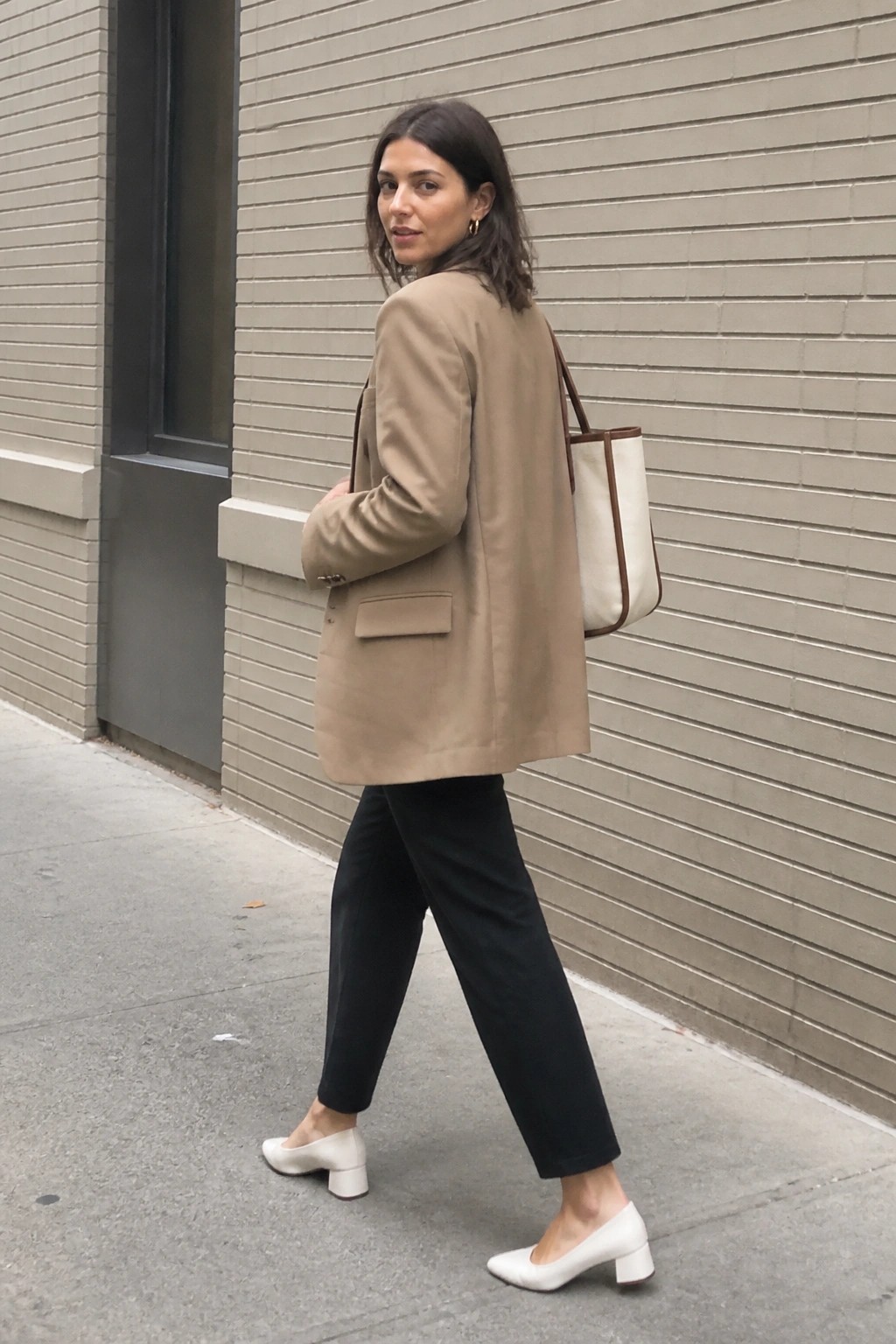 Woman in tan blazer and black pants carrying tote bag on sidewalk