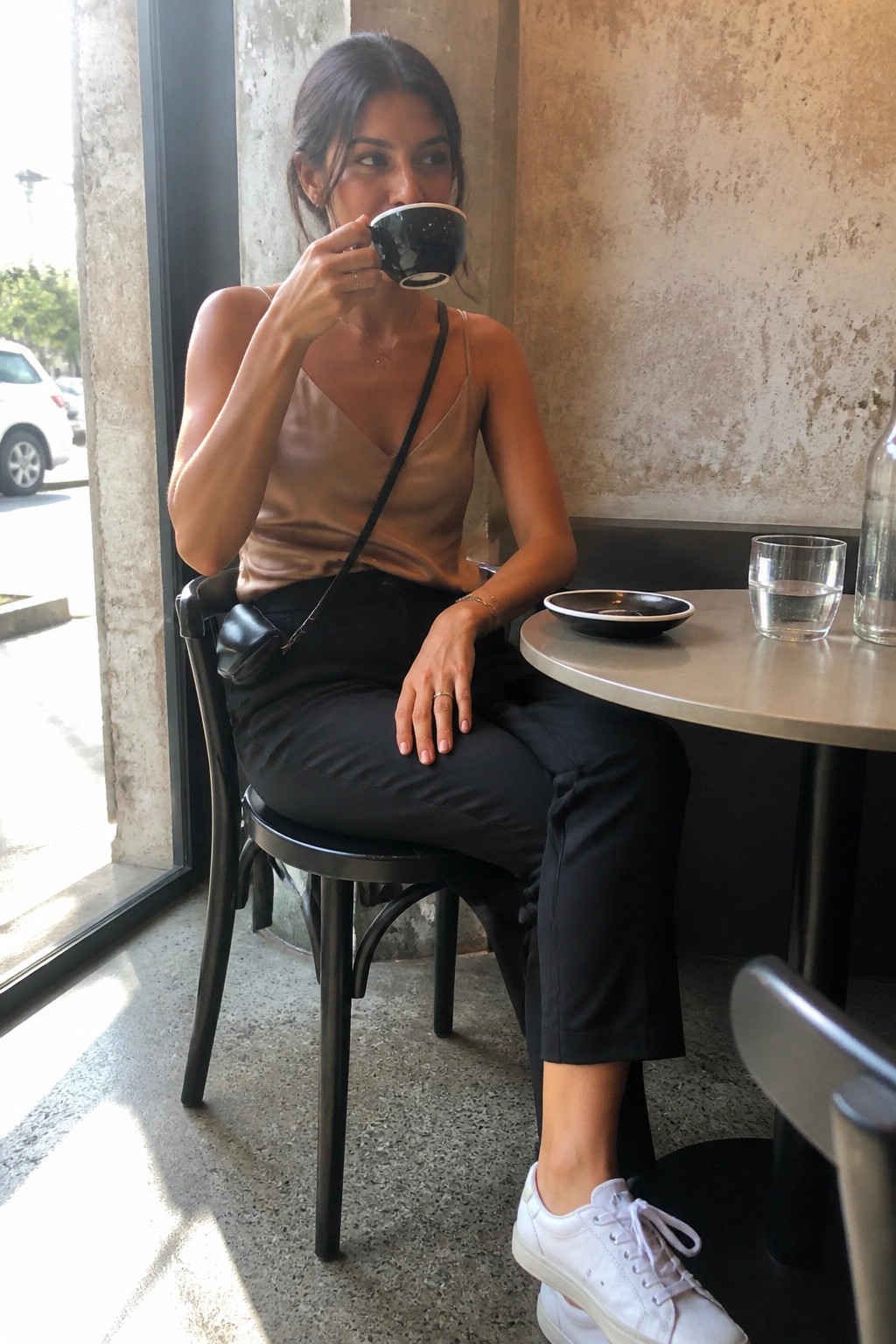 Woman in beige satin top sipping coffee at cafe table in black pants