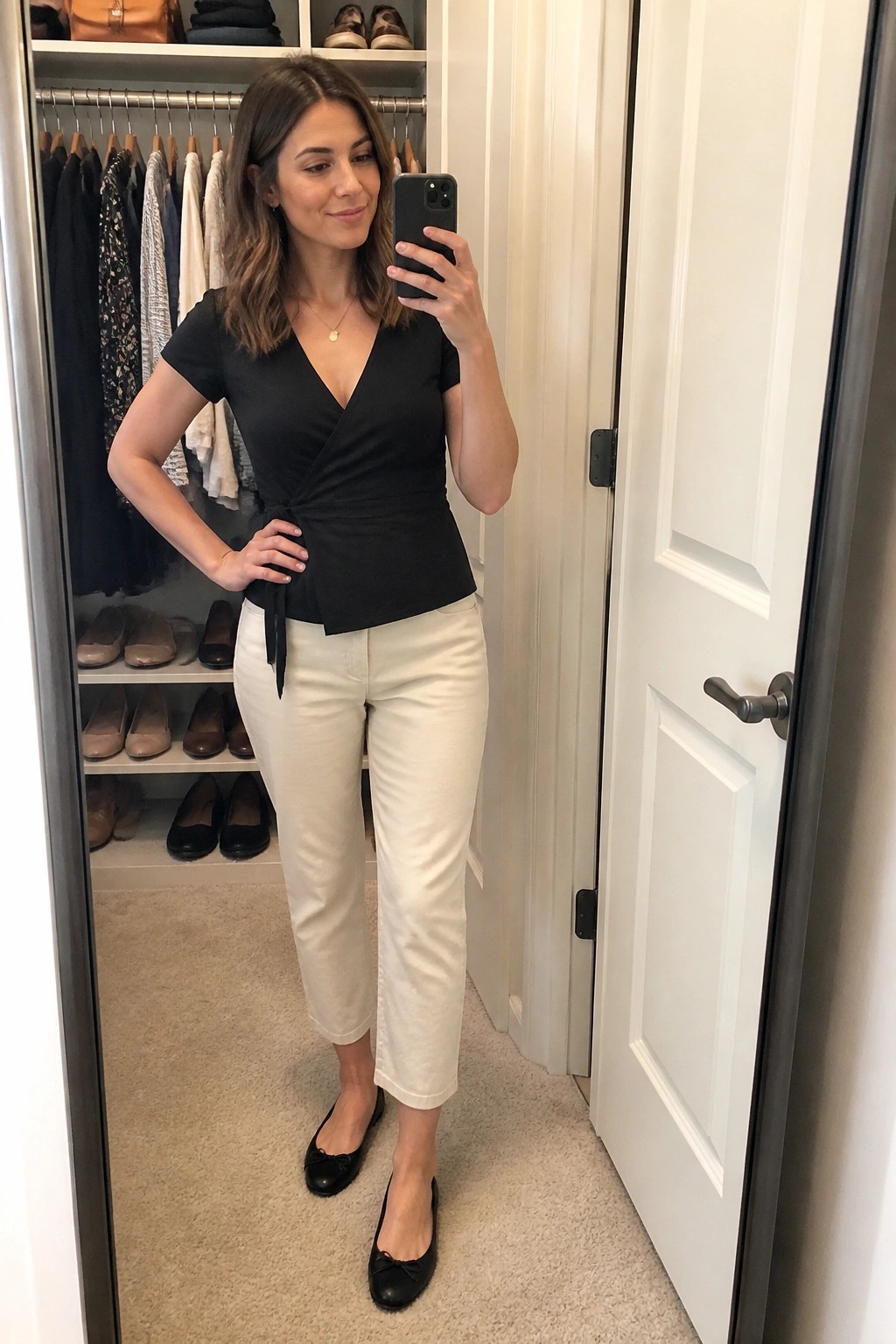 Woman in black wrap top and white capri pants taking mirror selfie in closet