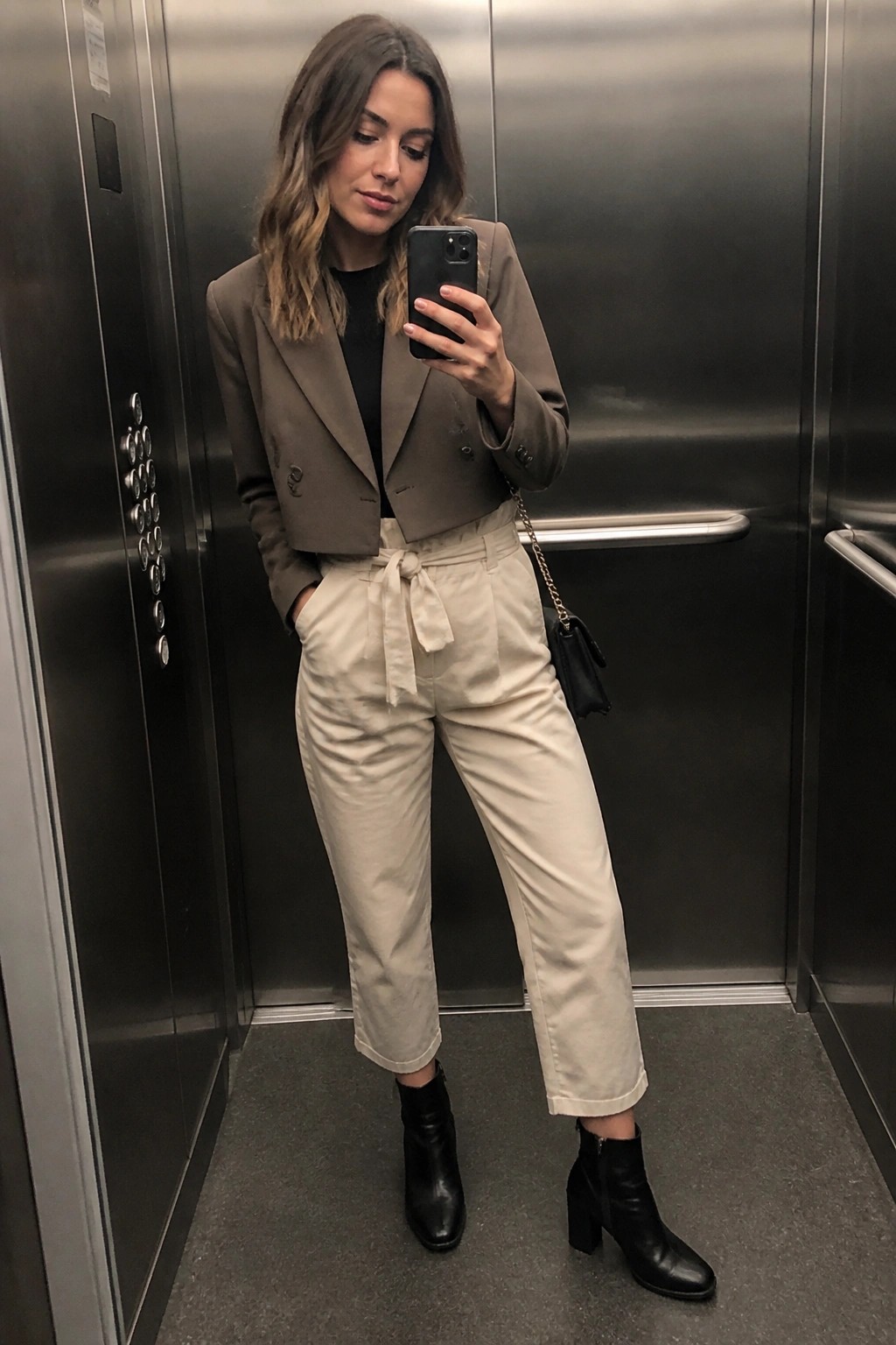 Woman in taupe cropped blazer and beige pants taking selfie in elevator