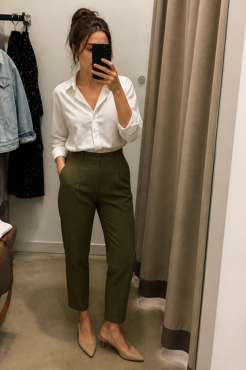 Woman in white shirt and olive pants taking mirror selfie in fitting room