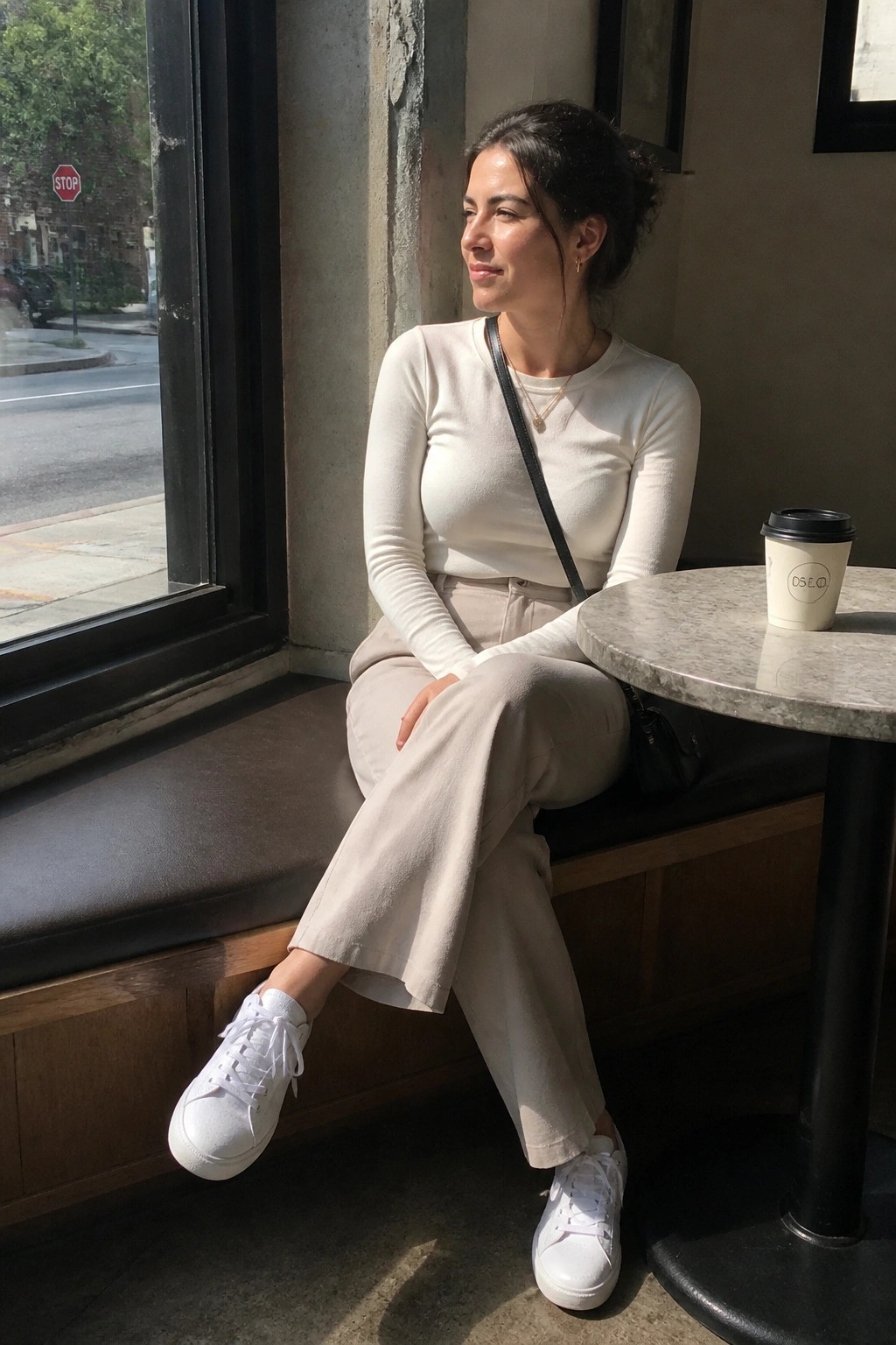 Woman in white top and beige pants seated by sunny cafe window with coffee