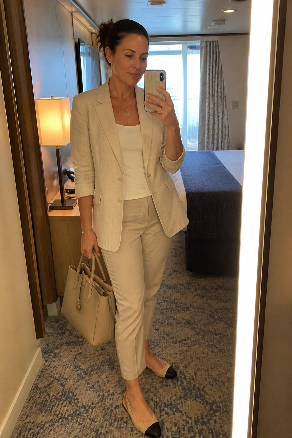 Woman in beige pantsuit taking mirror selfie while holding handbag in hotel room