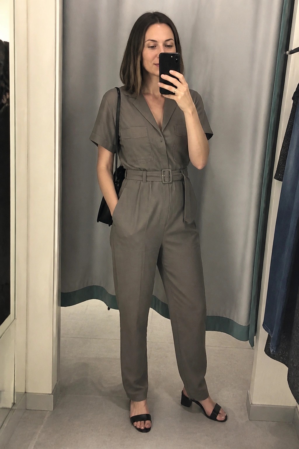 Woman in gray belted jumpsuit taking mirror selfie in fitting room.