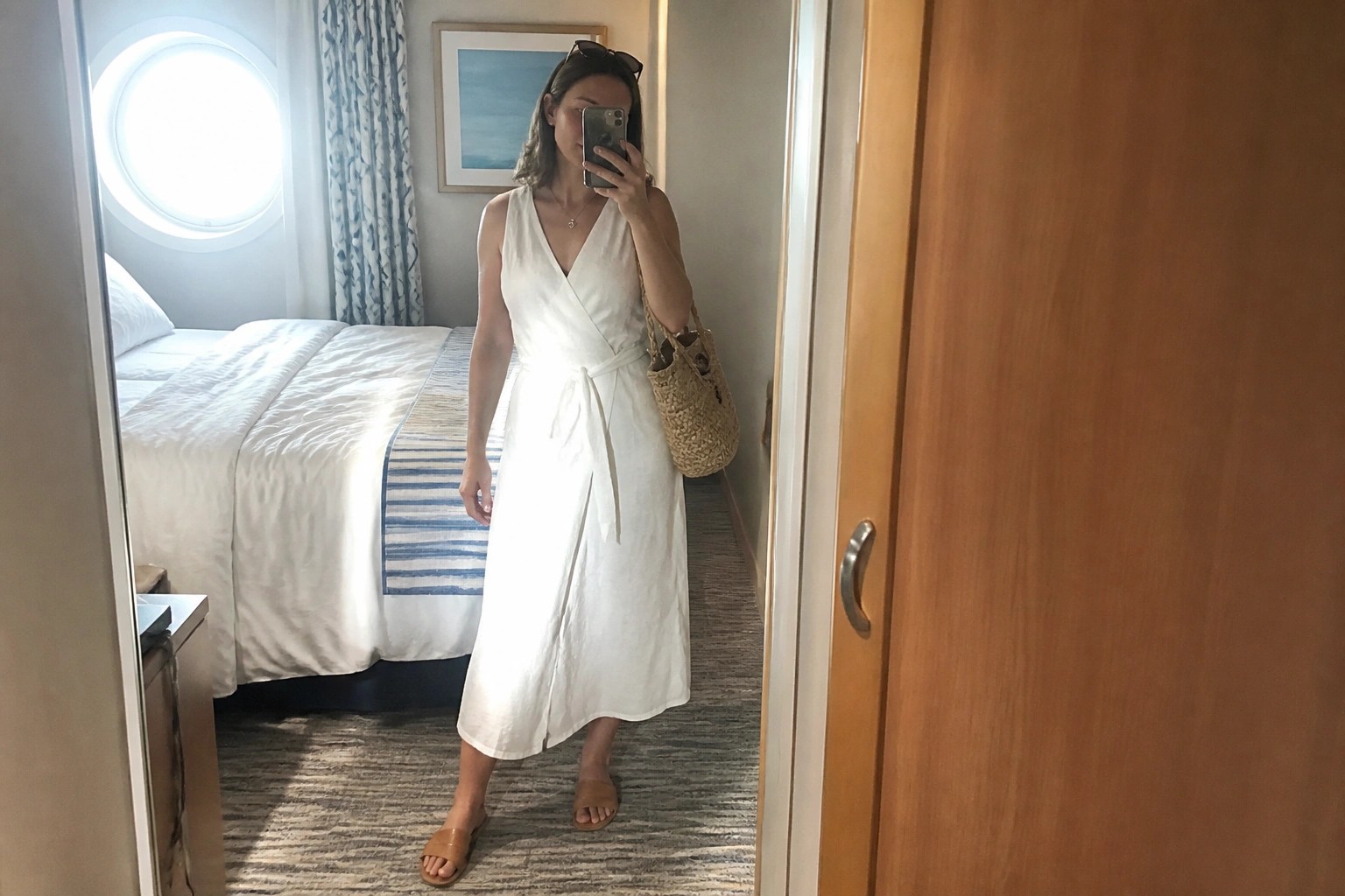 Woman wearing a white sleeveless wrap midi dress with waist tie belt, brown slide sandals, and a woven straw tote bag while taking a mirror selfie.