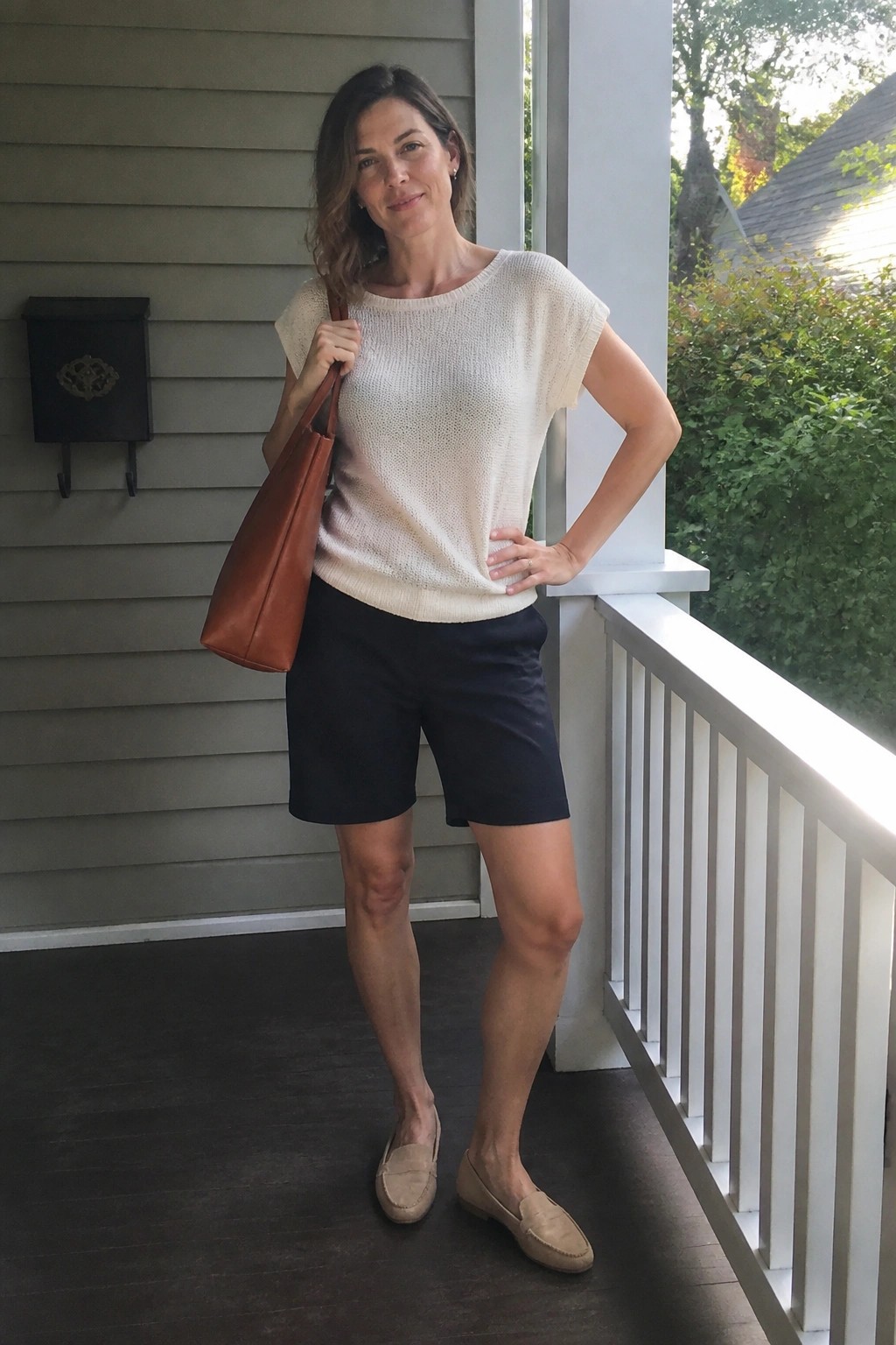 Woman in cream knit sweater and navy shorts holding brown leather tote on porch