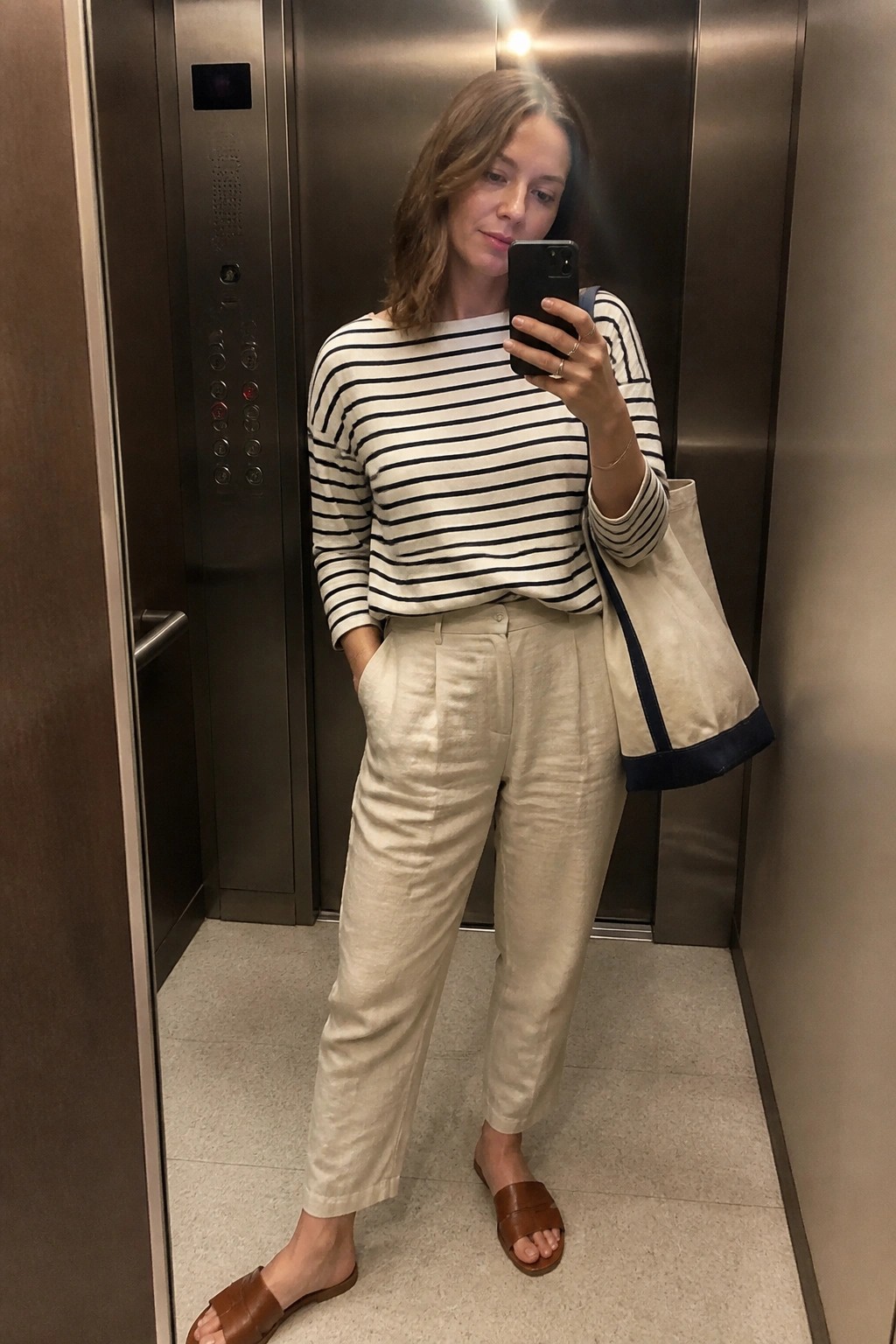 Woman in striped top and beige pants taking mirror selfie in elevator with tote bag.