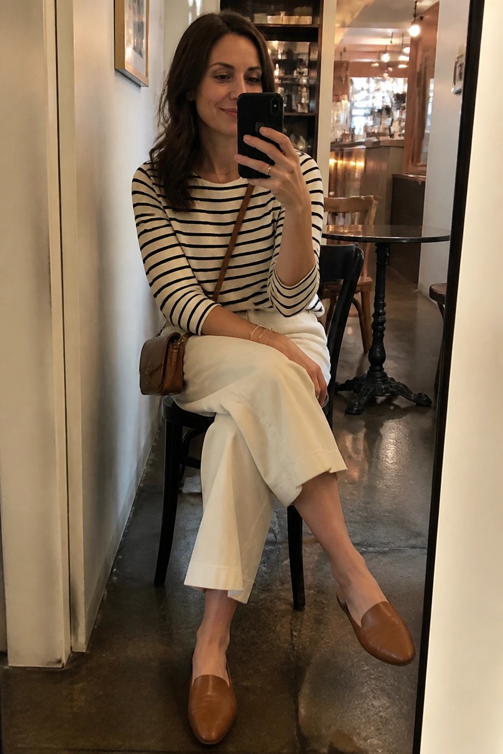 Woman in striped shirt and white pants taking mirror selfie in cafe
