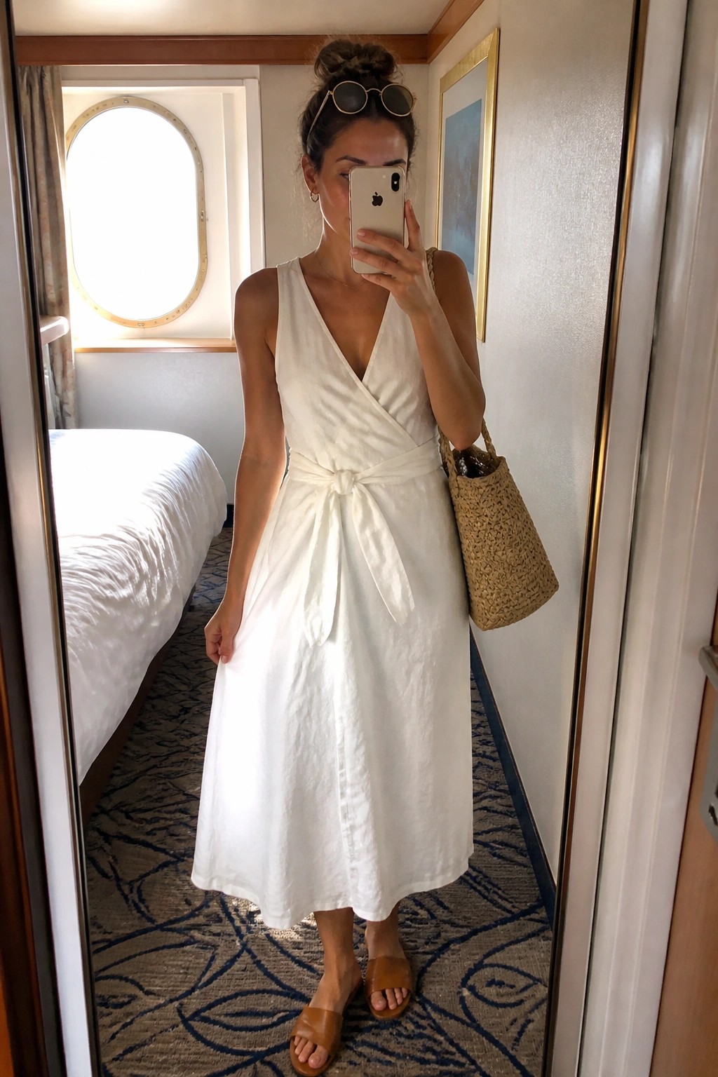 Woman wearing a white sleeveless wrap midi dress with waist tie belt, brown slide sandals, and a woven straw tote bag while taking a mirror selfie.