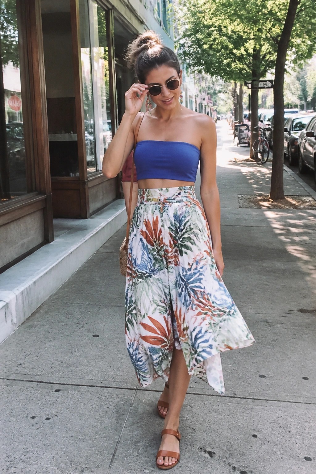 Woman in blue strapless top and floral skirt adjusts sunglasses on sidewalk