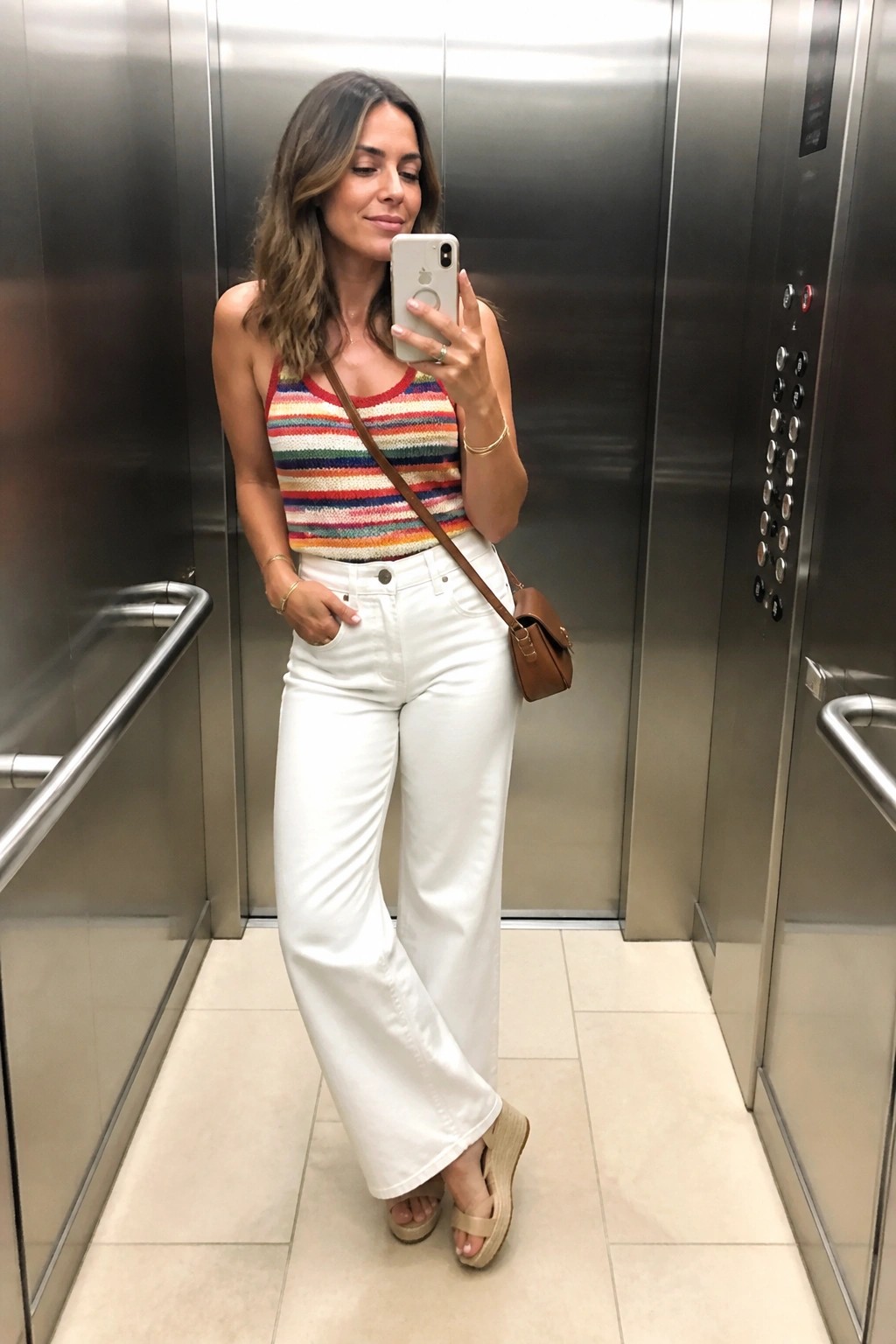 Woman in multicolored striped tank top and white wide-leg jeans taking elevator selfie