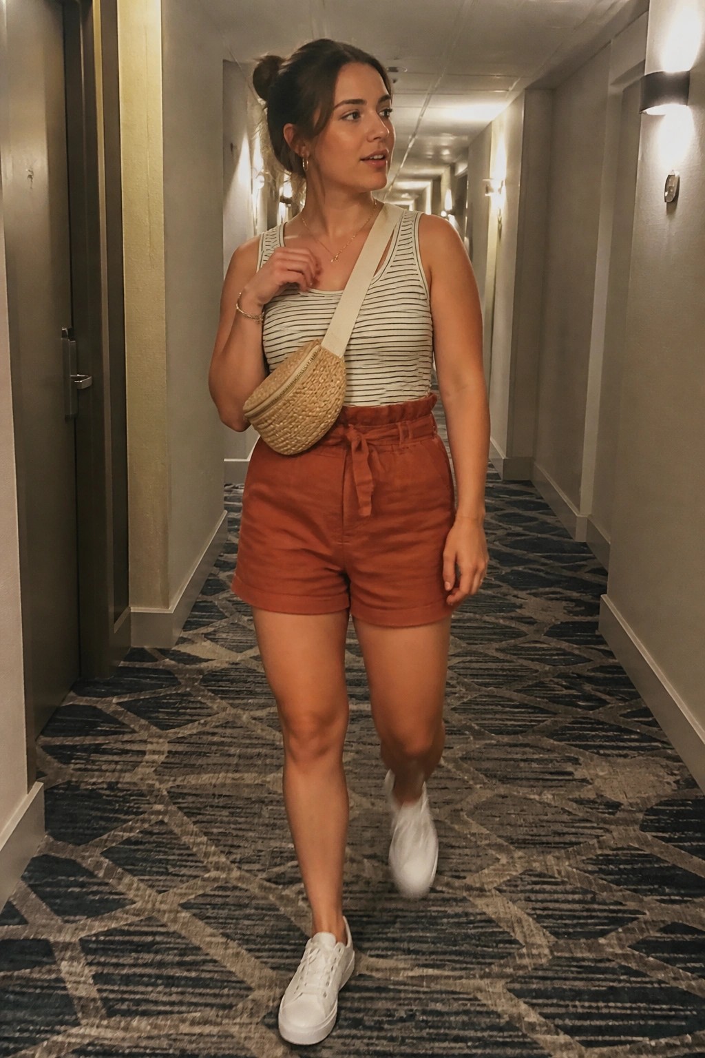 Woman in striped tank top and rust shorts with woven bag in hallway