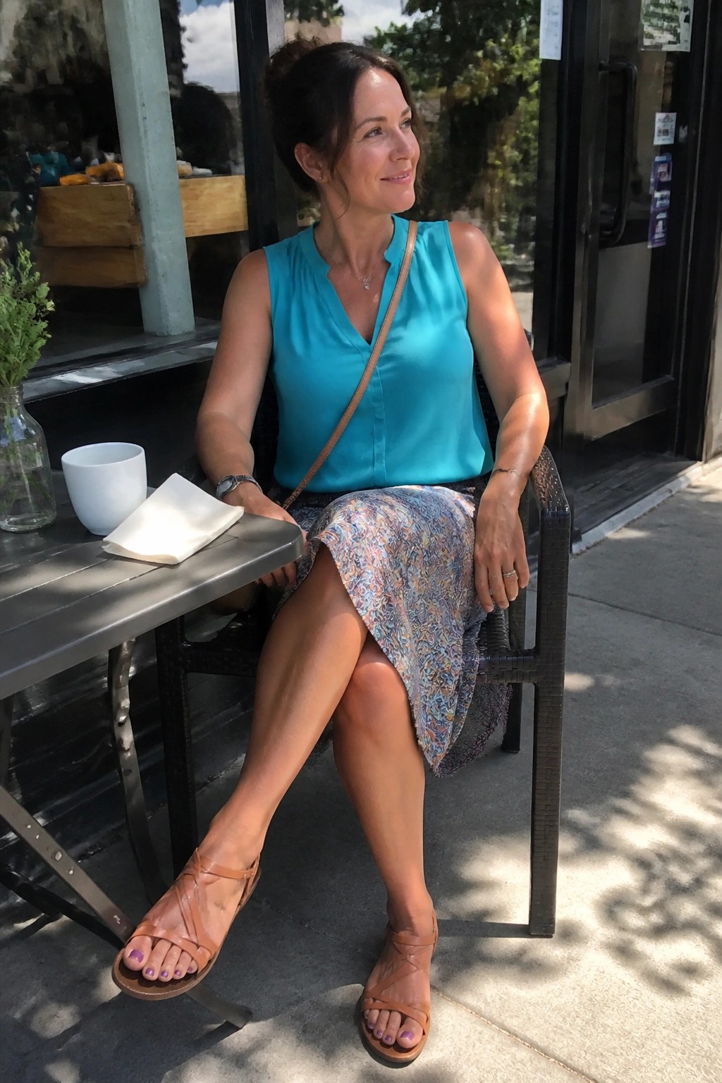 Woman in turquoise top and patterned skirt seated at sunny outdoor cafe table