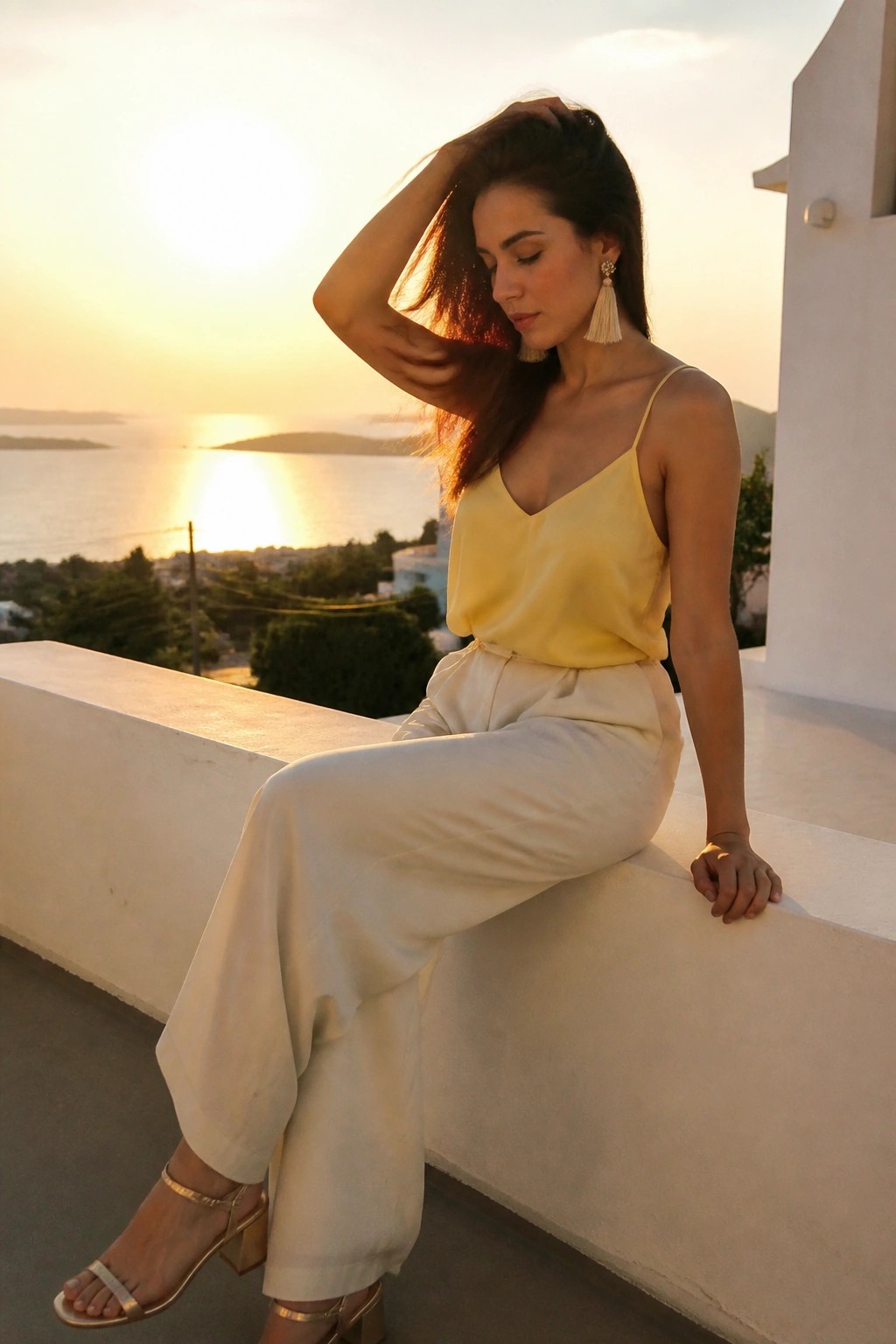 Woman in yellow camisole and white pants on ledge overlooking sunset sea