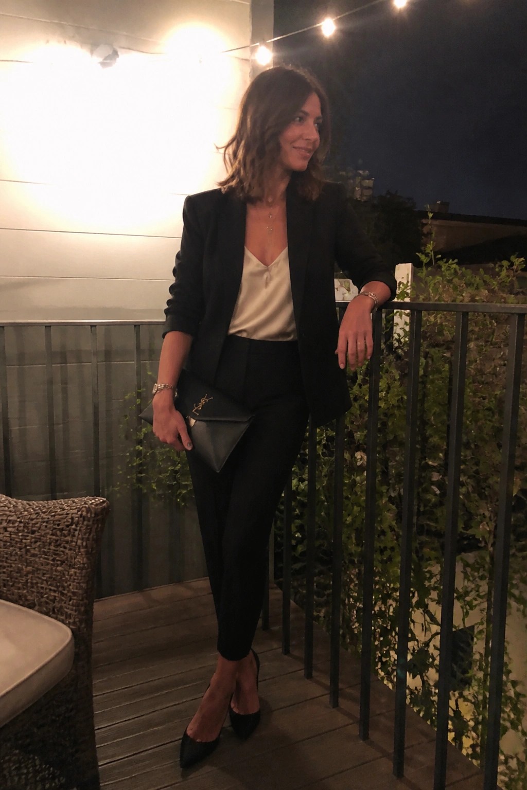 Woman in black blazer and pants holding YSL clutch on nighttime balcony