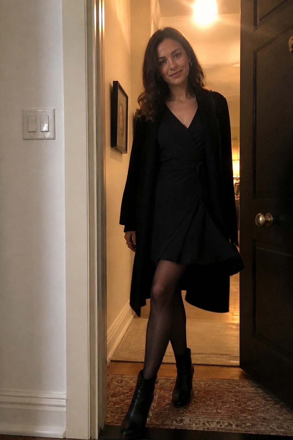 Woman wearing a black wrap dress under a long black coat with sheer black tights and black ankle boots in a doorway setting.