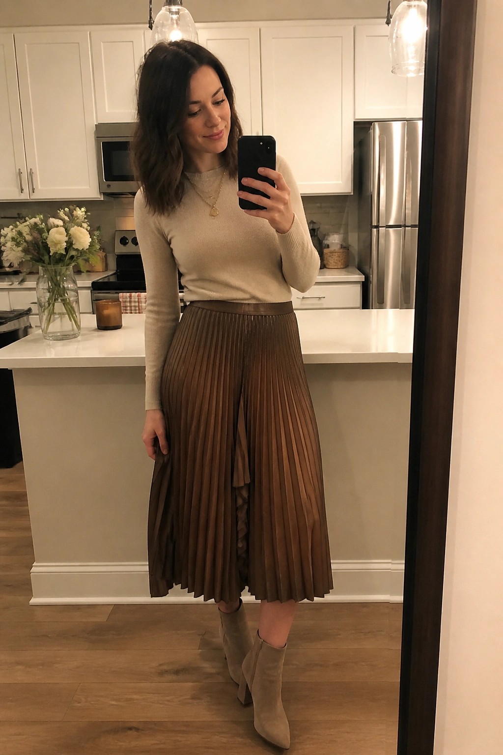 Woman in cream knit sweater brown pleated midi skirt and beige ankle boots posing for mirror selfie in kitchen setting