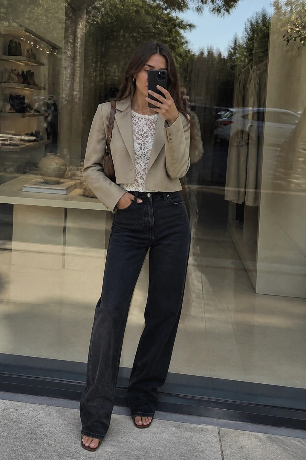 Woman in a cropped beige blazer over a white lace top, paired with dark wide-leg jeans, brown shoulder bag, and brown slide sandals, taking a mirror selfie indoors.