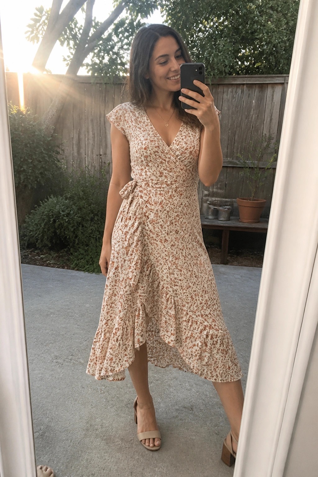 Woman in floral wrap dress takes mirror selfie outdoors in sunlight.