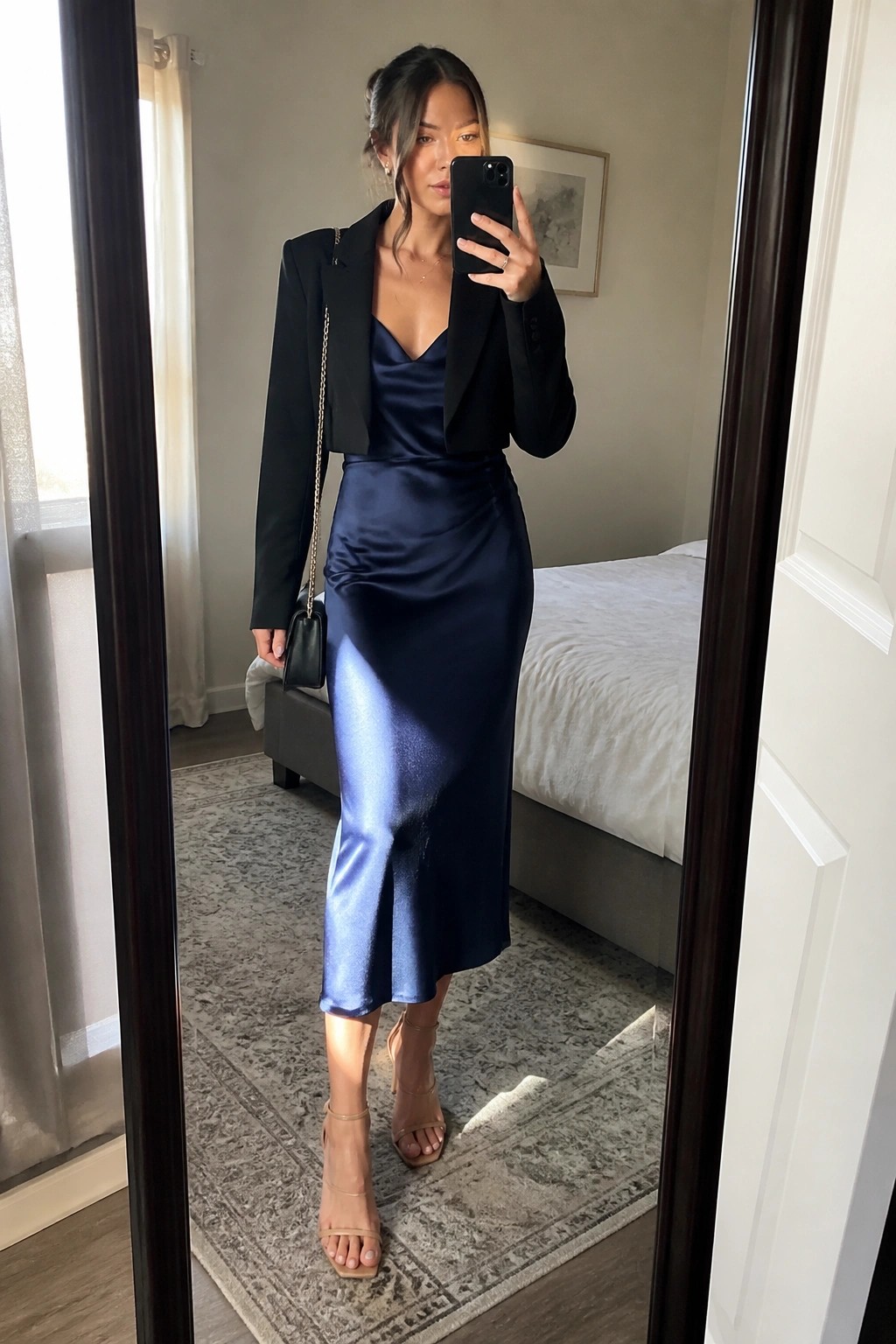 Woman in mirror selfie wearing navy blue satin midi slip dress under cropped black blazer, with nude strappy heels and small black handbag on gold chain strap.