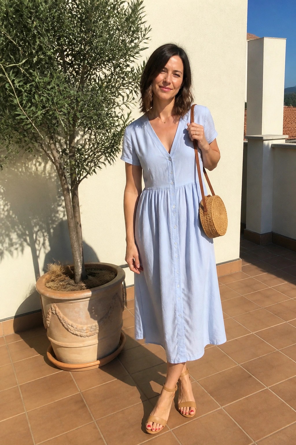 Woman in light blue button front midi dress carrying round woven straw bag and wearing beige block heel sandals.