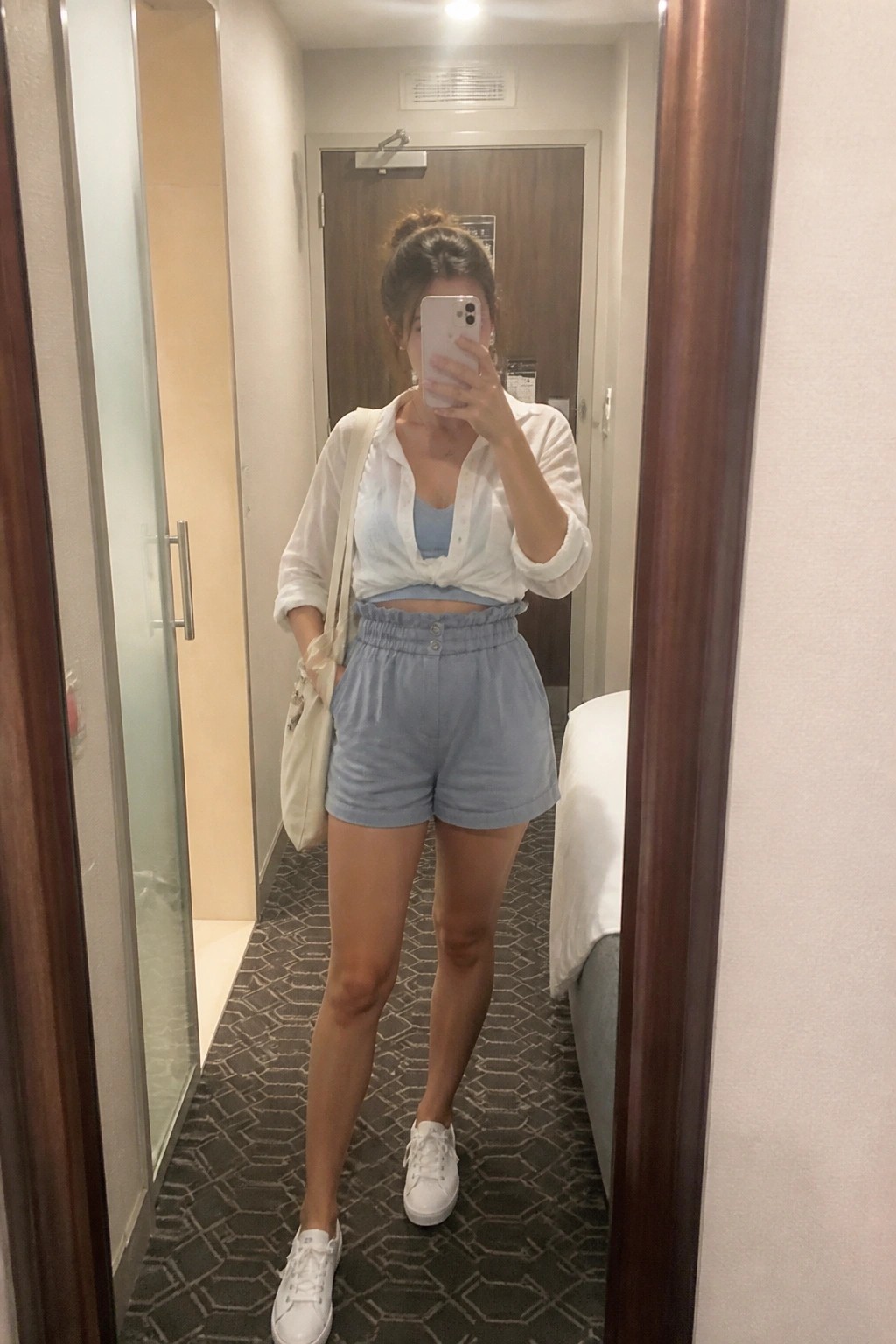 Woman in tied white shirt and blue shorts taking mirror selfie in hotel hallway