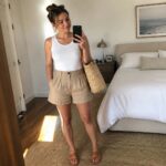 Woman in white ribbed tank top and beige shorts holding a straw tote while wearing brown sandals and taking a mirror selfie.