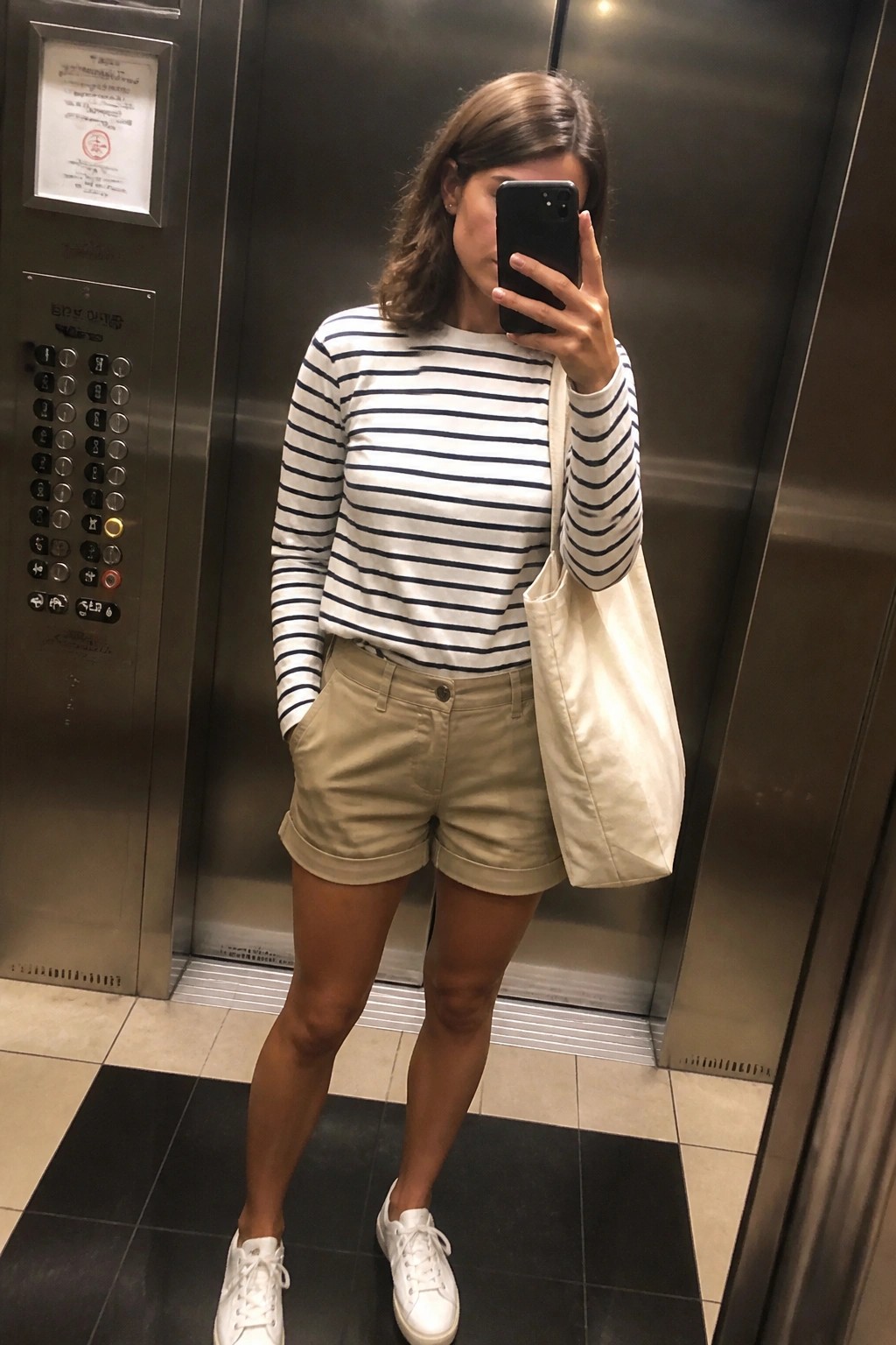 Woman in striped shirt and khaki shorts taking selfie in elevator with tote bag