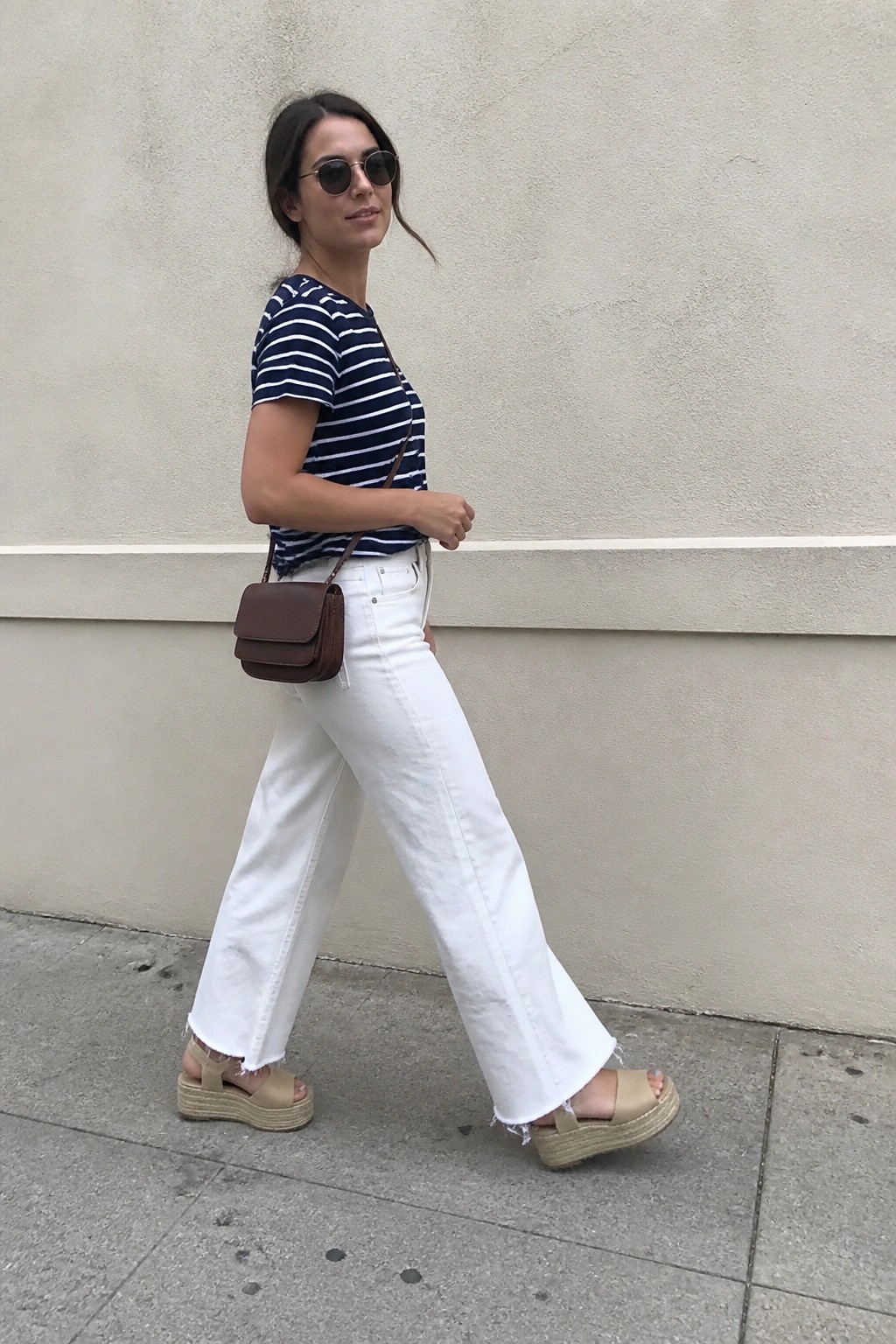 Woman in navy and white striped short sleeve tee, white wide leg frayed jeans, beige platform espadrilles, and brown crossbody bag walking on pavement.