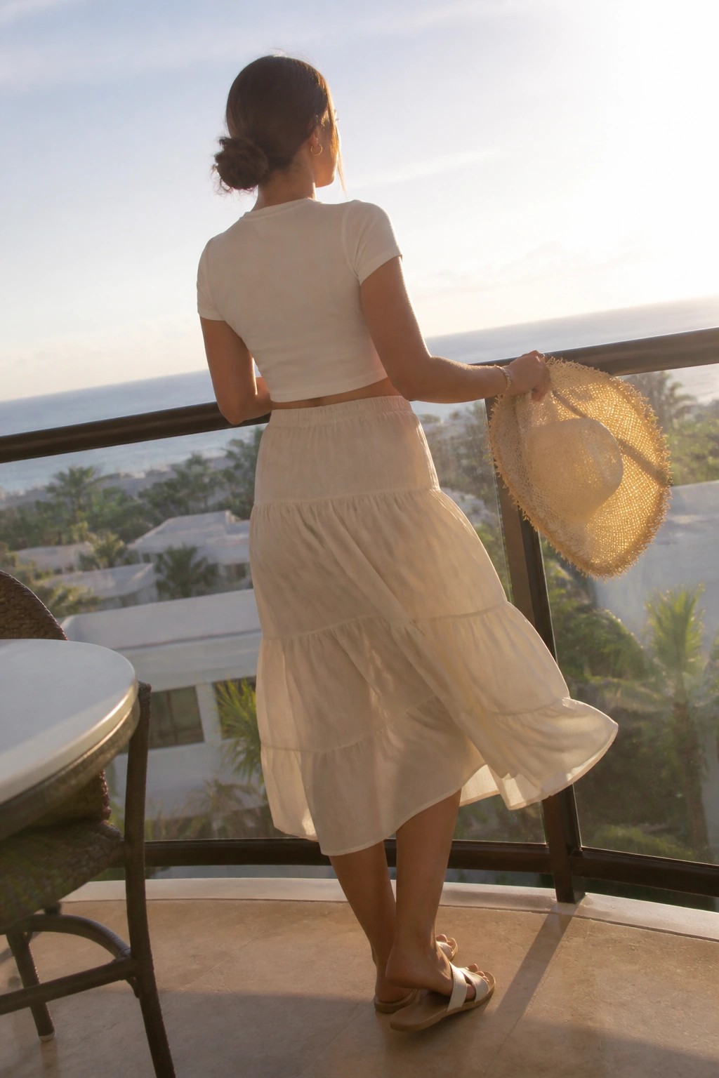 Woman in white tiered skirt holding straw hat on ocean-view balcony