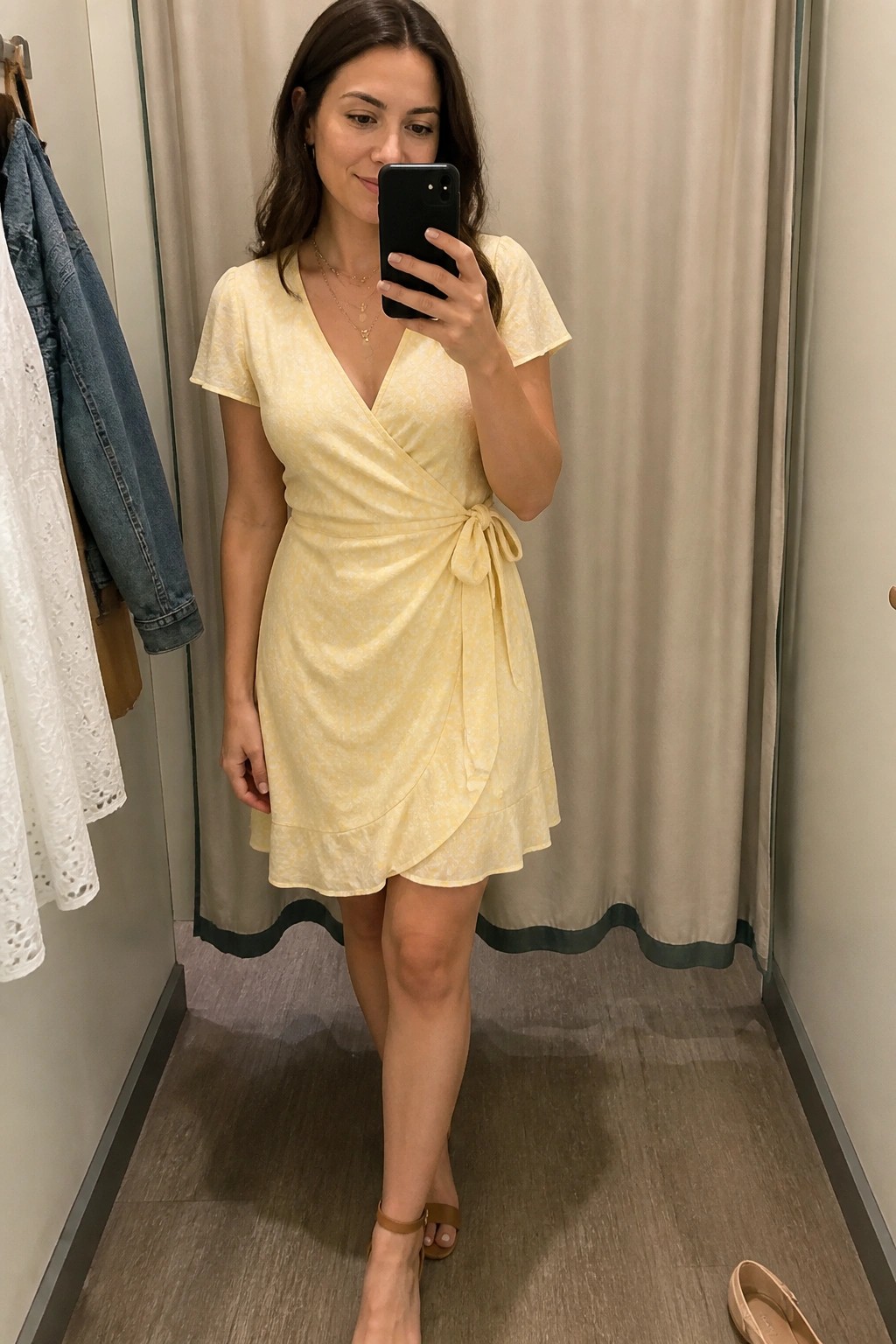 Woman in pale yellow floral wrap dress taking mirror selfie in fitting room