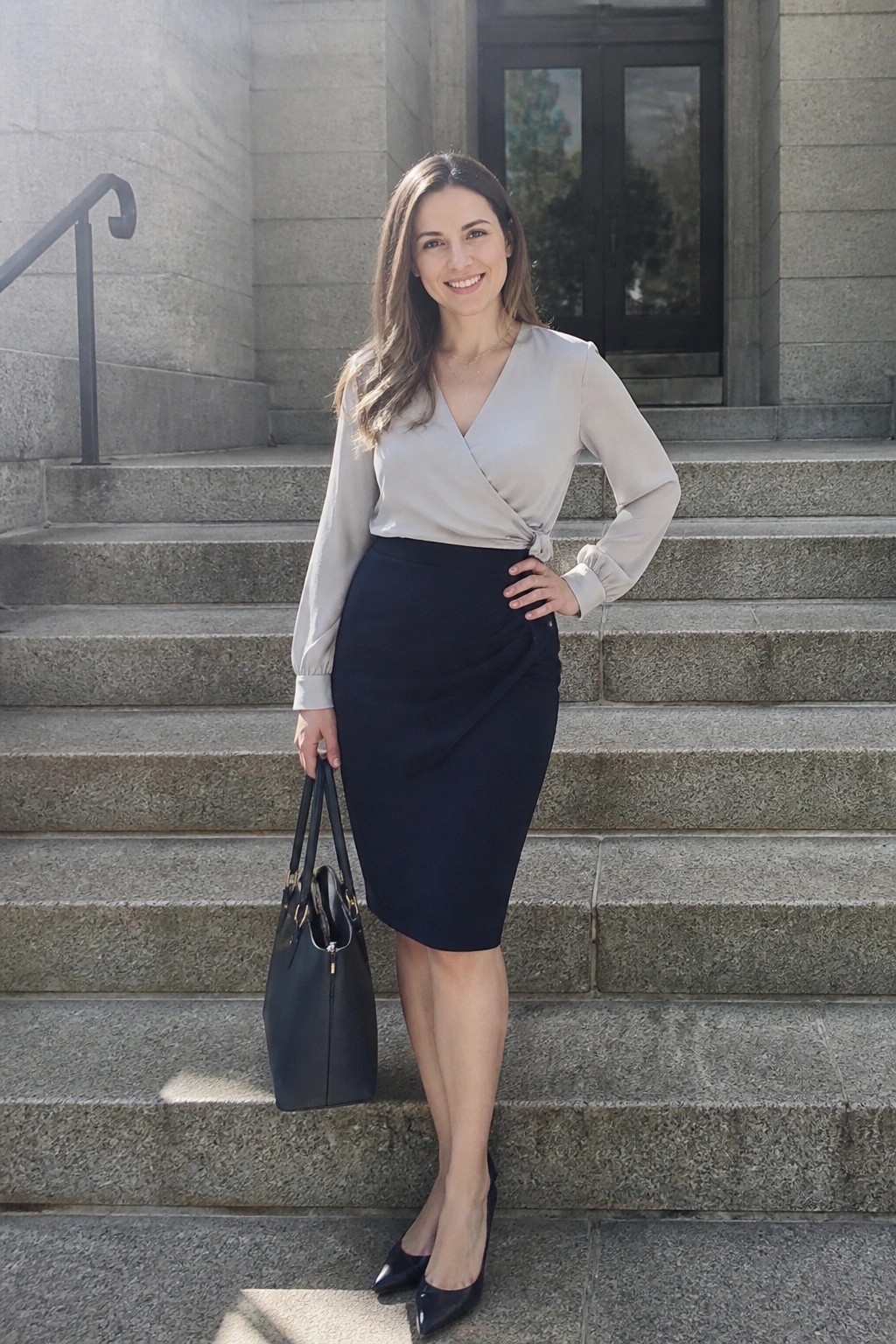 Smiling woman in beige wrap blouse and navy skirt holding black bag on steps