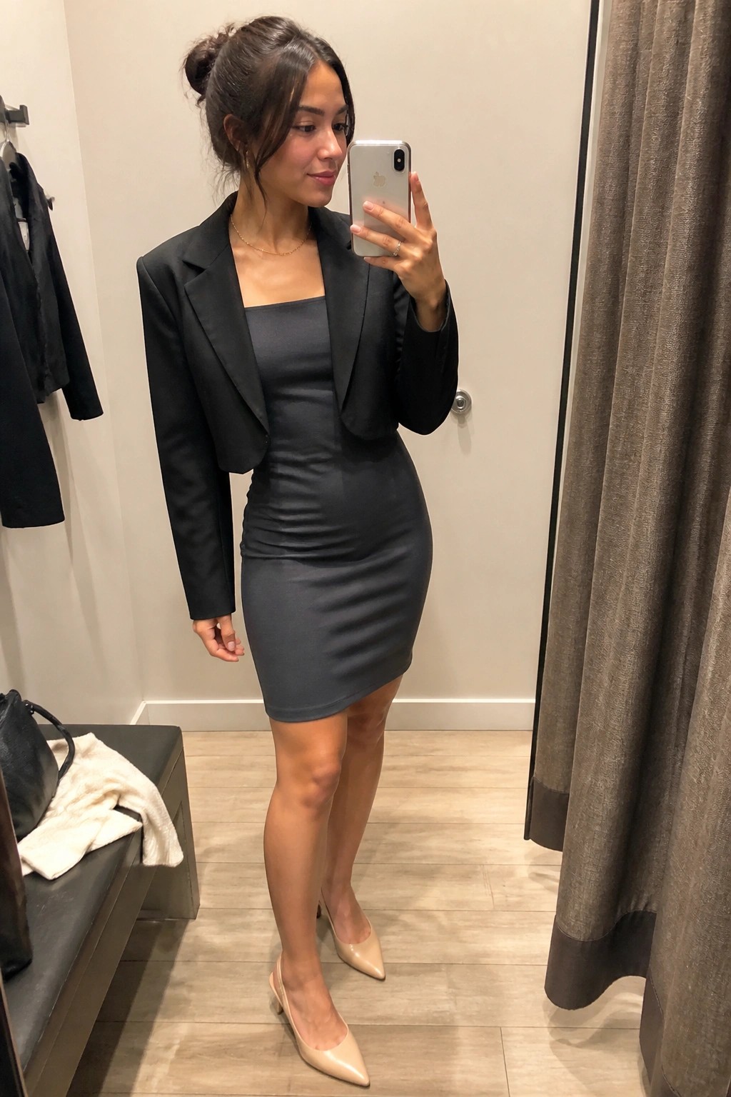 Woman in black cropped blazer and gray mini dress taking mirror selfie in fitting room