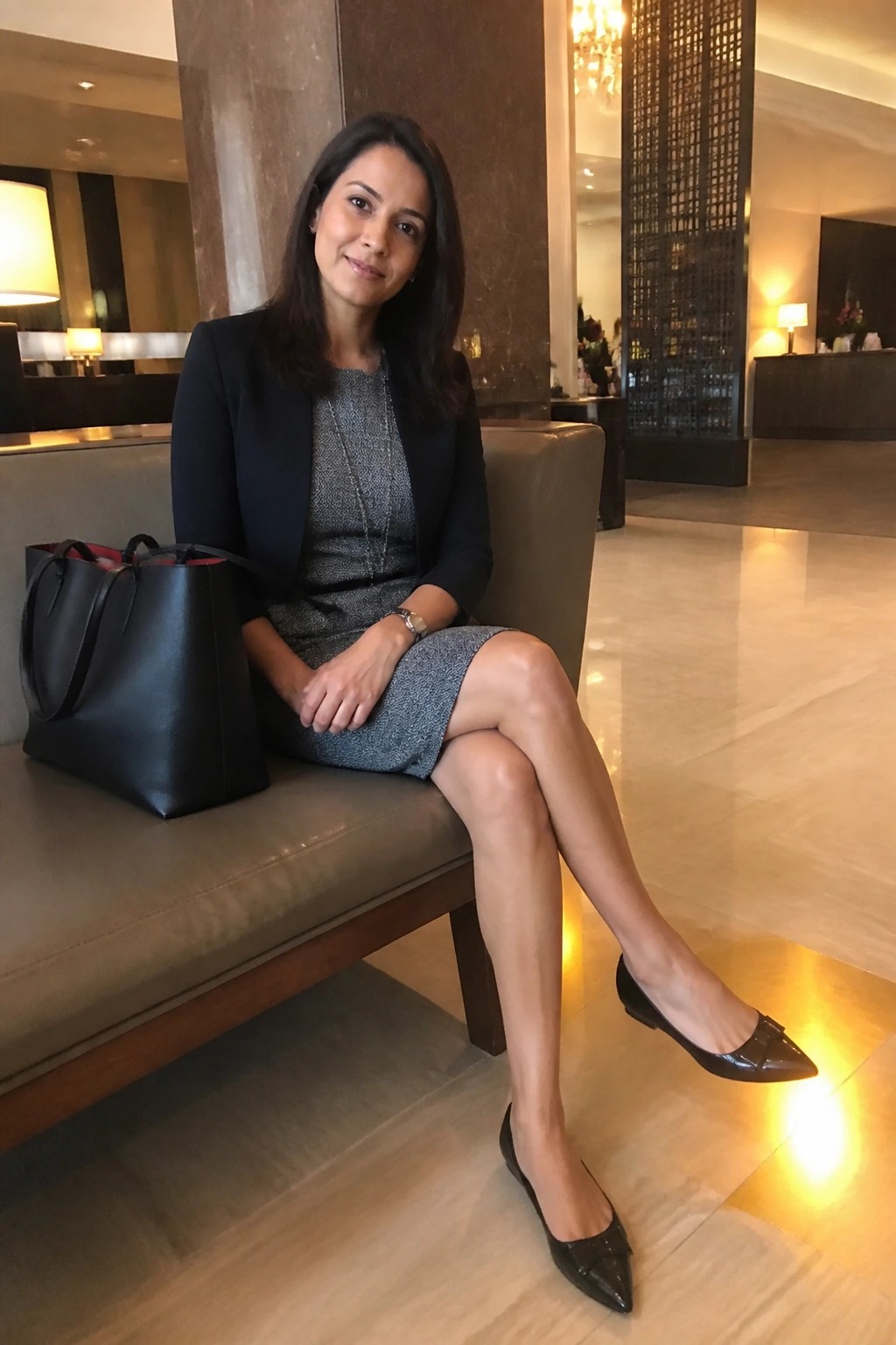 Woman in black blazer and gray dress seated on bench beside black handbag in lobby