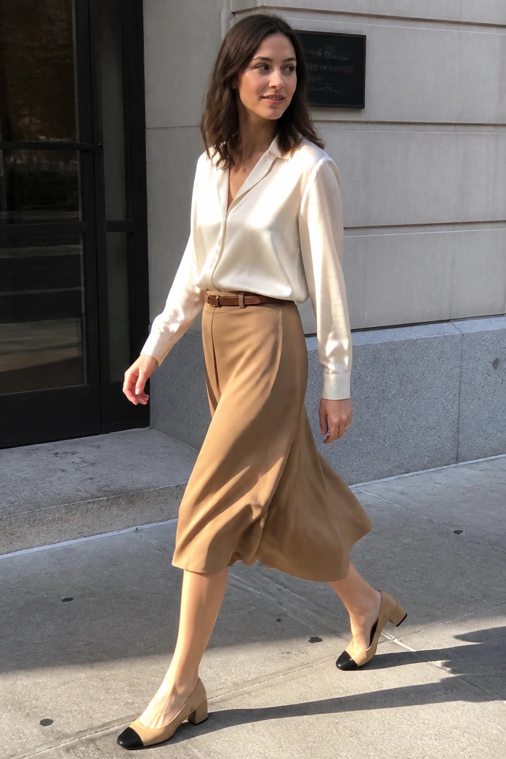 Woman in white satin blouse and tan skirt walking on urban sidewalk in two-tone shoes