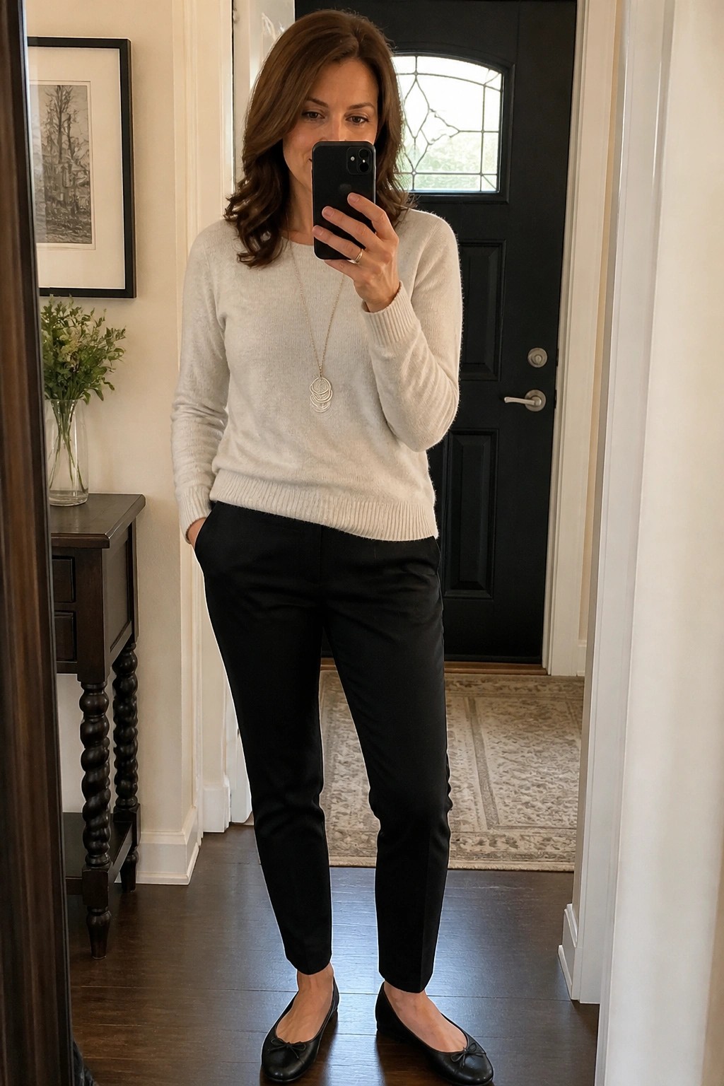 Woman in cream sweater and black pants taking mirror selfie indoors