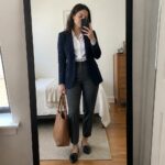 Woman wearing a navy blazer over a white button-up shirt with gray tailored trousers, black pointed loafers, and a brown leather tote bag in a mirror selfie.