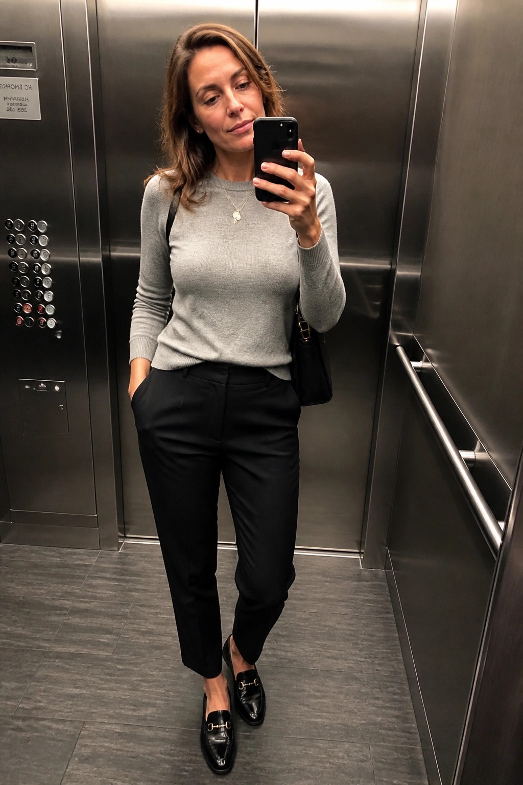 Woman wearing a fitted gray knit sweater with black tailored trousers and black patent loafers while holding a phone for a mirror selfie in an elevator.