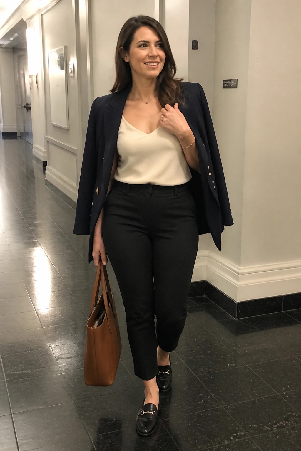 Woman in navy blazer, cream top, black pants holding brown tote in hallway