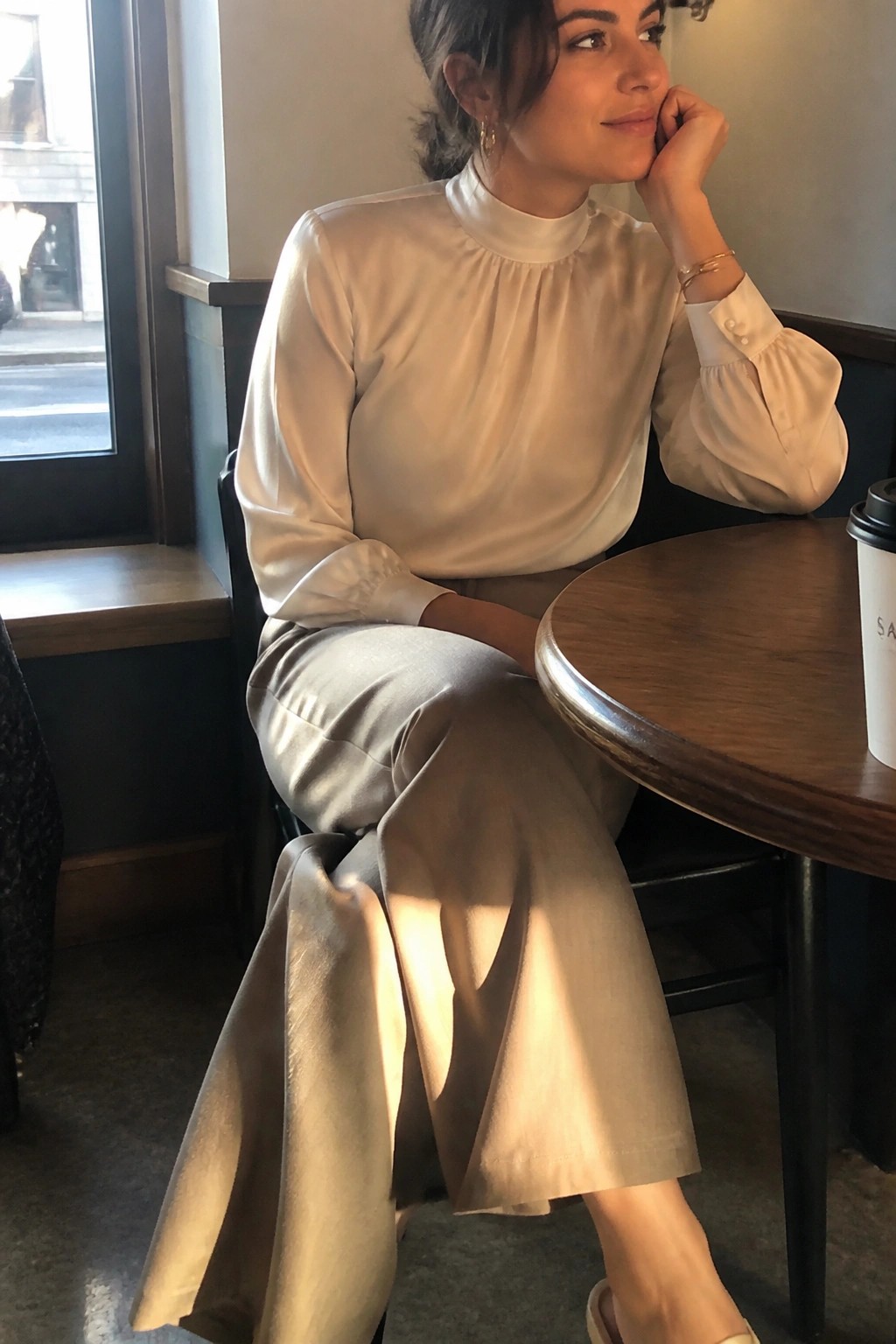 Woman in cream satin blouse and beige skirt at sunlit cafe table with coffee