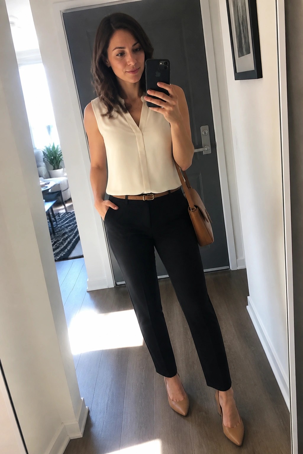 Woman in cream sleeveless V-neck blouse tucked into black trousers with brown belt, brown pointed-toe heels, and brown shoulder bag taking a mirror selfie.