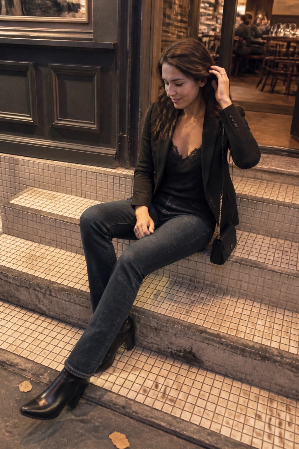 Woman in black blazer and jeans sits on tiled steps, hand in hair.