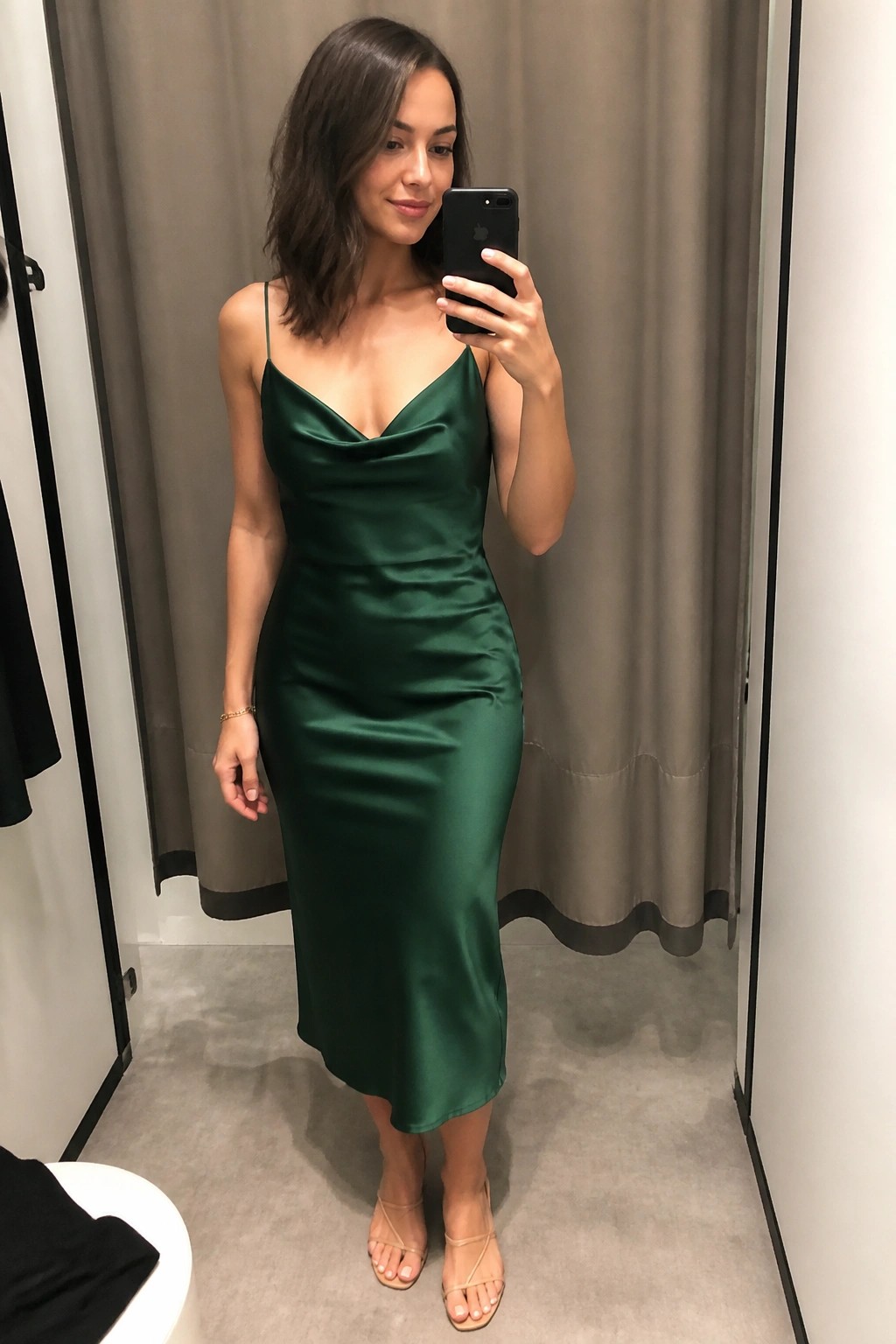 Woman in emerald green satin dress taking mirror selfie in fitting room